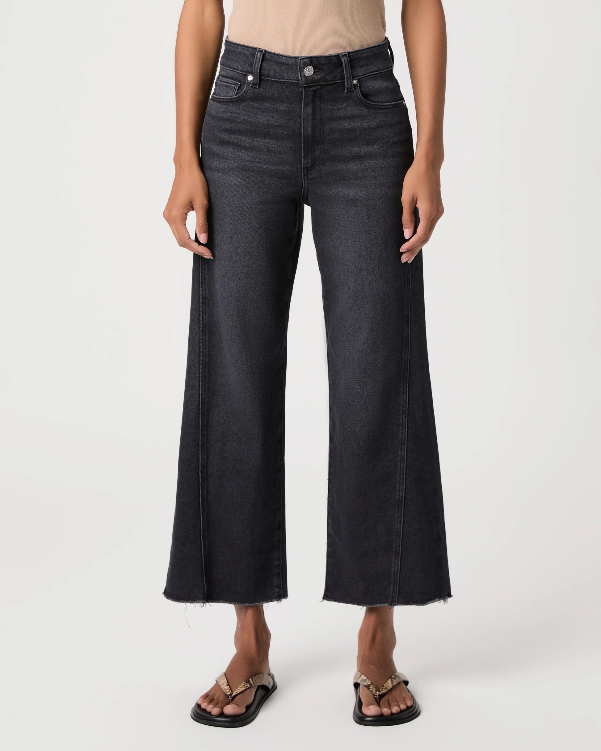 A model wearing a pair of dark charcoal gray wide-leg cropped jeans with a raw hem, paired with black slide sandals, shown against a plain light gray studio background.