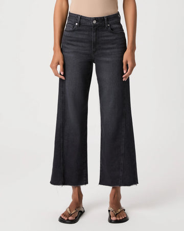 A model wearing a pair of dark charcoal gray wide-leg cropped jeans with a raw hem, paired with black slide sandals, shown against a plain light gray studio background.