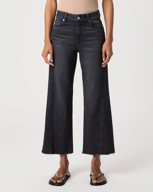 A model wearing a pair of dark charcoal gray wide-leg cropped jeans with a raw hem, paired with black slide sandals, shown against a plain light gray studio background.