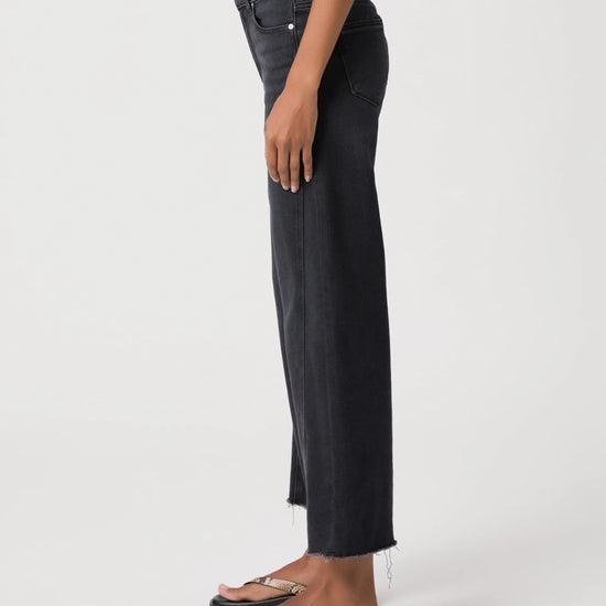 A side-profile view of a person wearing dark charcoal gray cropped jeans with a raw hem and black slide sandals, photographed against a clean white studio background.
