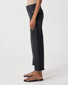 A side-profile view of a person wearing dark charcoal gray cropped jeans with a raw hem and black slide sandals, photographed against a clean white studio background.