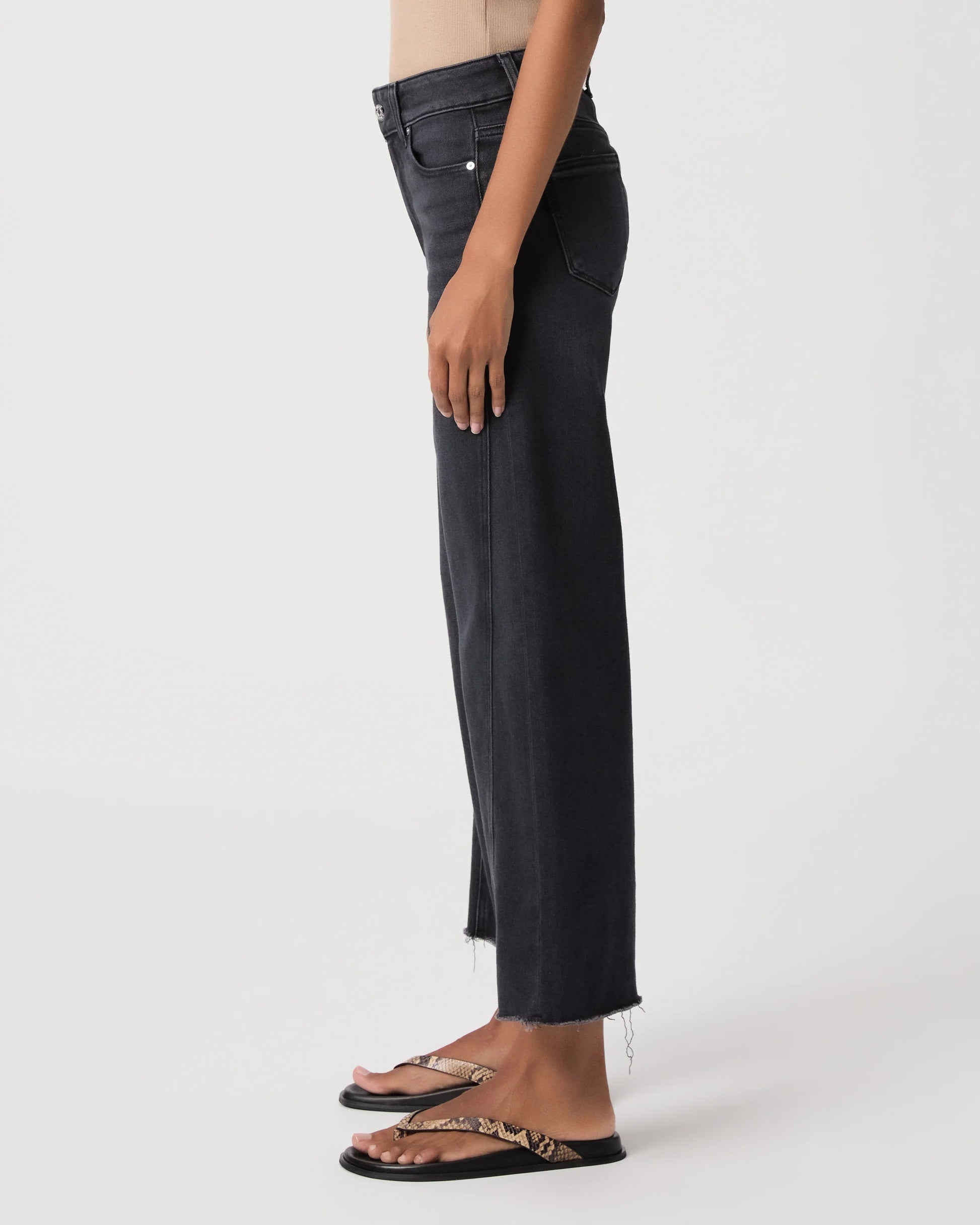 A side-profile view of a person wearing dark charcoal gray cropped jeans with a raw hem and black slide sandals, photographed against a clean white studio background.
