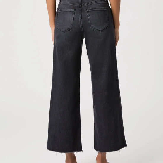 Rear view of a model wearing dark charcoal gray wide-leg cropped jeans with a frayed hem and black sandals against a light gray studio background.