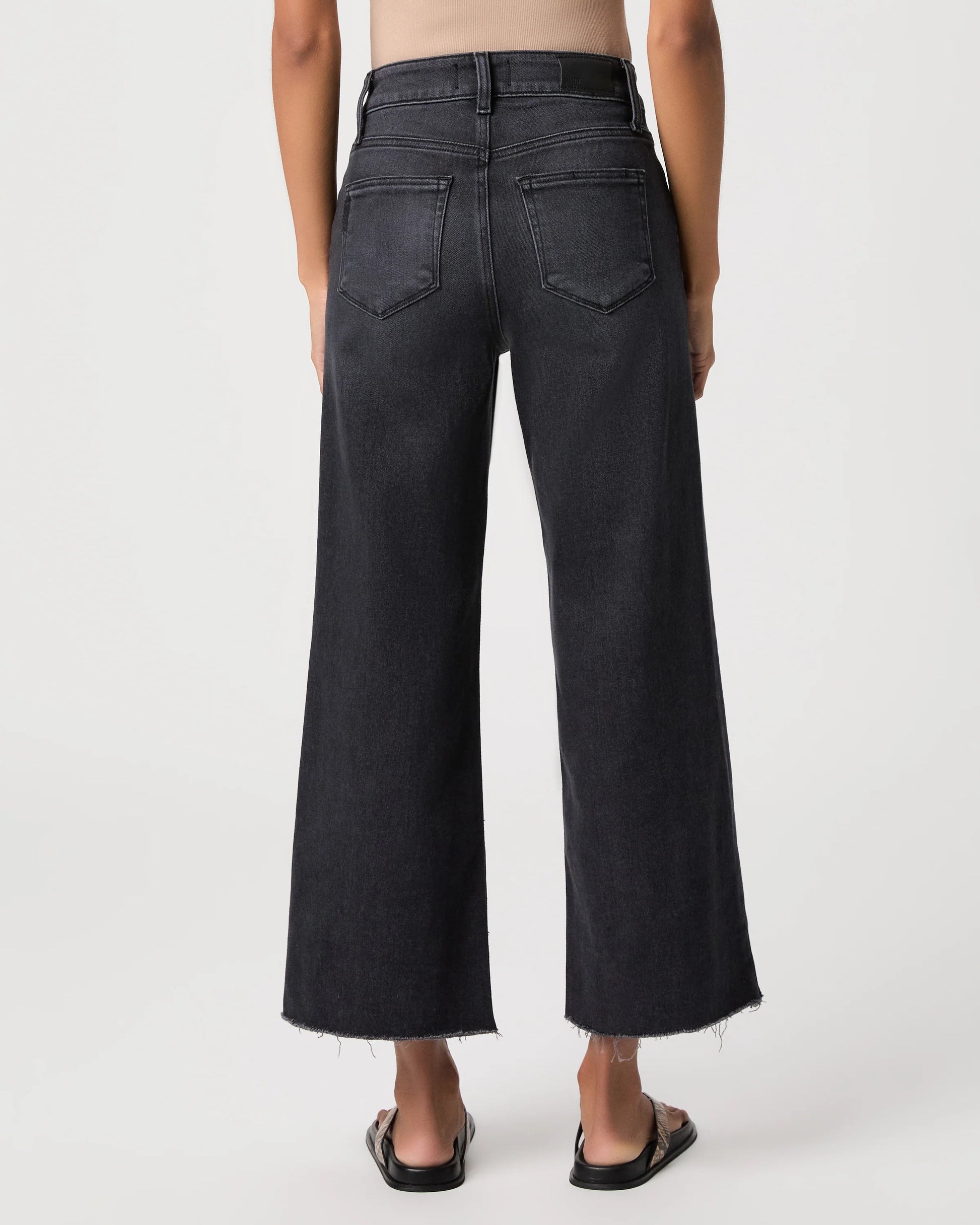 Rear view of a model wearing dark charcoal gray wide-leg cropped jeans with a frayed hem and black sandals against a light gray studio background.