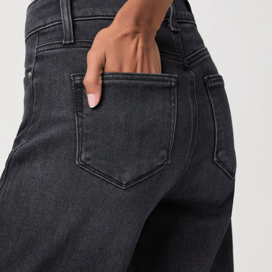 Close-up of a person wearing dark charcoal gray jeans, showing the back pocket and hand in the pocket.