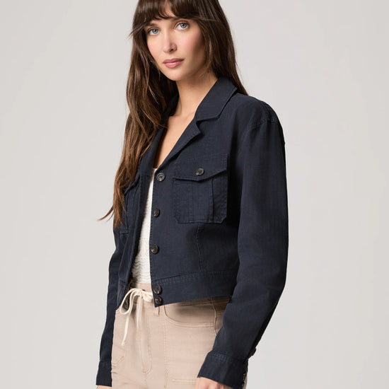 Woman wearing a navy jacket over a white top and beige pants on a light gray background