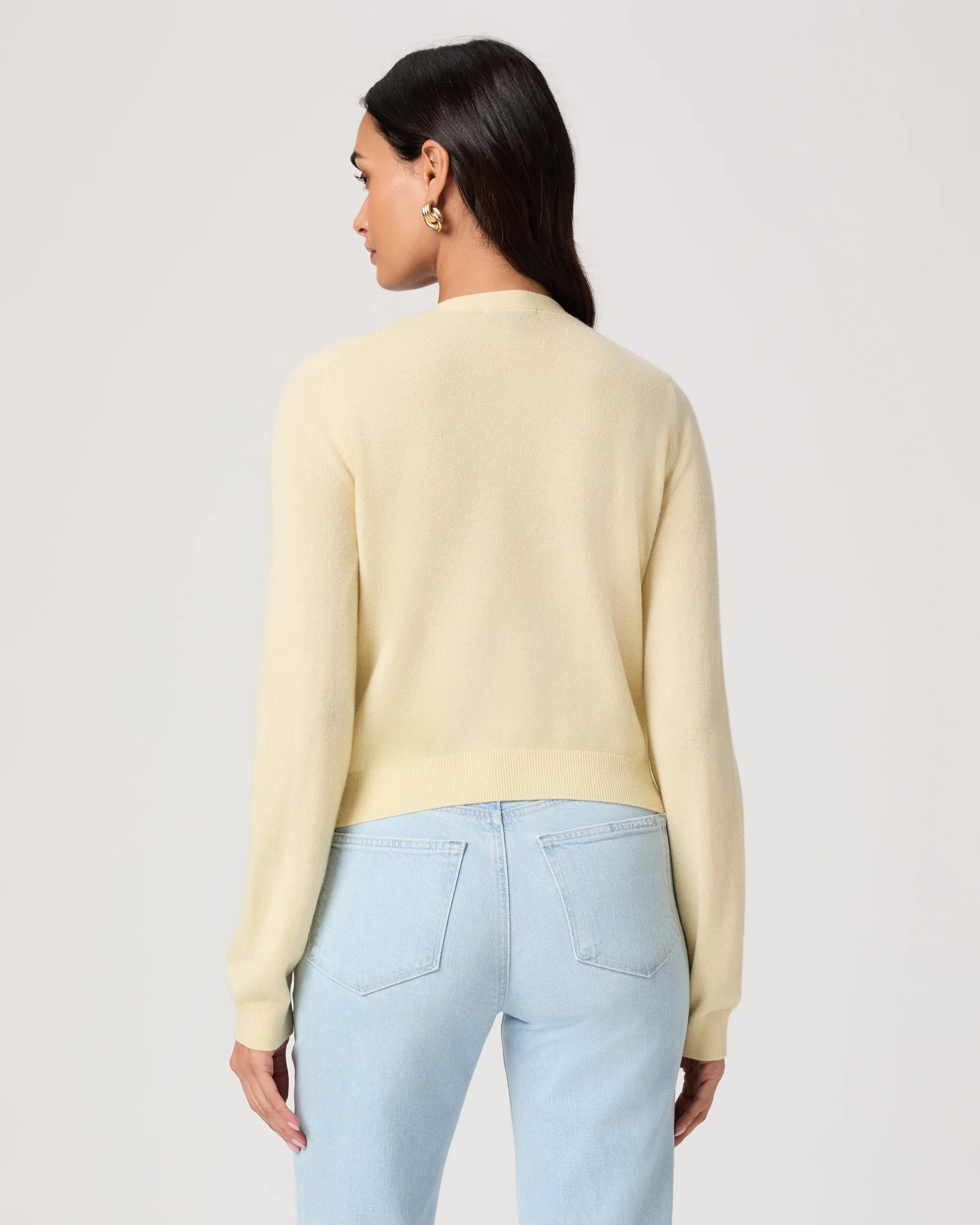 Woman wearing a light yellow cardigan by Paige and light blue jeans on a white background rear view