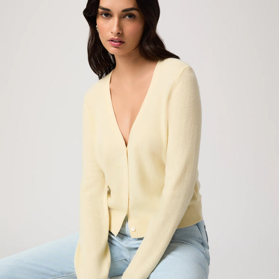 Woman wearing a light yellow cardigan by Paige and light blue jeans on a white background, sat down