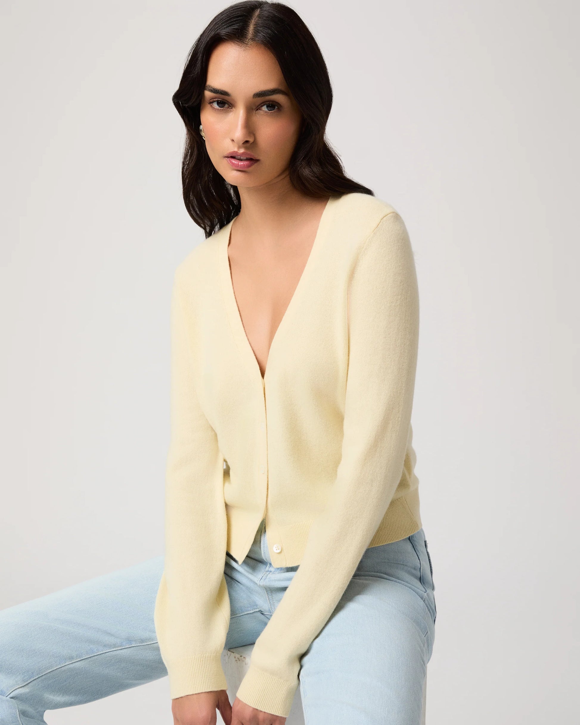 Woman wearing a light yellow cardigan by Paige and light blue jeans on a white background, sat down