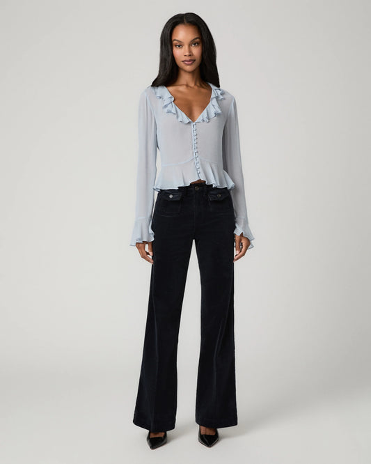 A woman modeling a light blue long-sleeve blouse with ruffle trim and dark wide-leg jeans against a plain light gray studio background.
