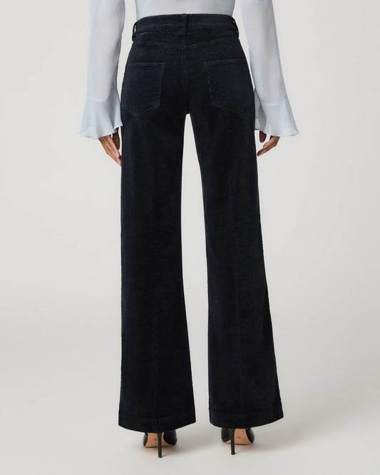 A model viewed from the back wearing high-waisted, wide-leg dark navy corduroy trousers, a white long-sleeve blouse, and black heels against a light gray studio background.