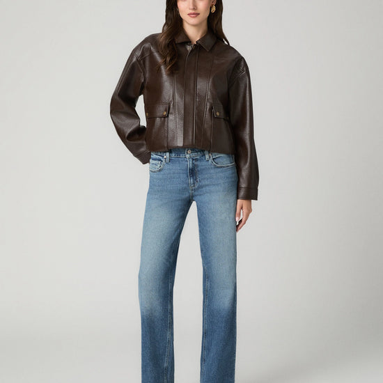 A model stands against a light gray studio background wearing a cropped dark brown leather jacket, blue straight-leg jeans, and black pointed-toe heels.