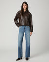 A model stands against a light gray studio background wearing a cropped dark brown leather jacket, blue straight-leg jeans, and black pointed-toe heels.