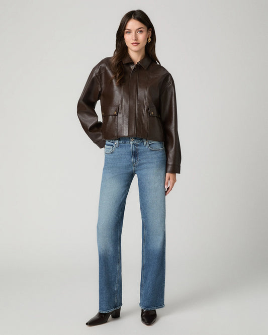 A model stands against a light gray studio background wearing a cropped dark brown leather jacket, blue straight-leg jeans, and black pointed-toe heels.