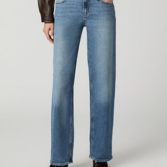 A model wearing a pair of medium-wash blue straight-leg jeans, styled with a brown leather jacket and dark brown pointed-toe boots, against a light gray studio background.