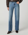 A model wearing a pair of medium-wash blue straight-leg jeans, styled with a brown leather jacket and dark brown pointed-toe boots, against a light gray studio background.