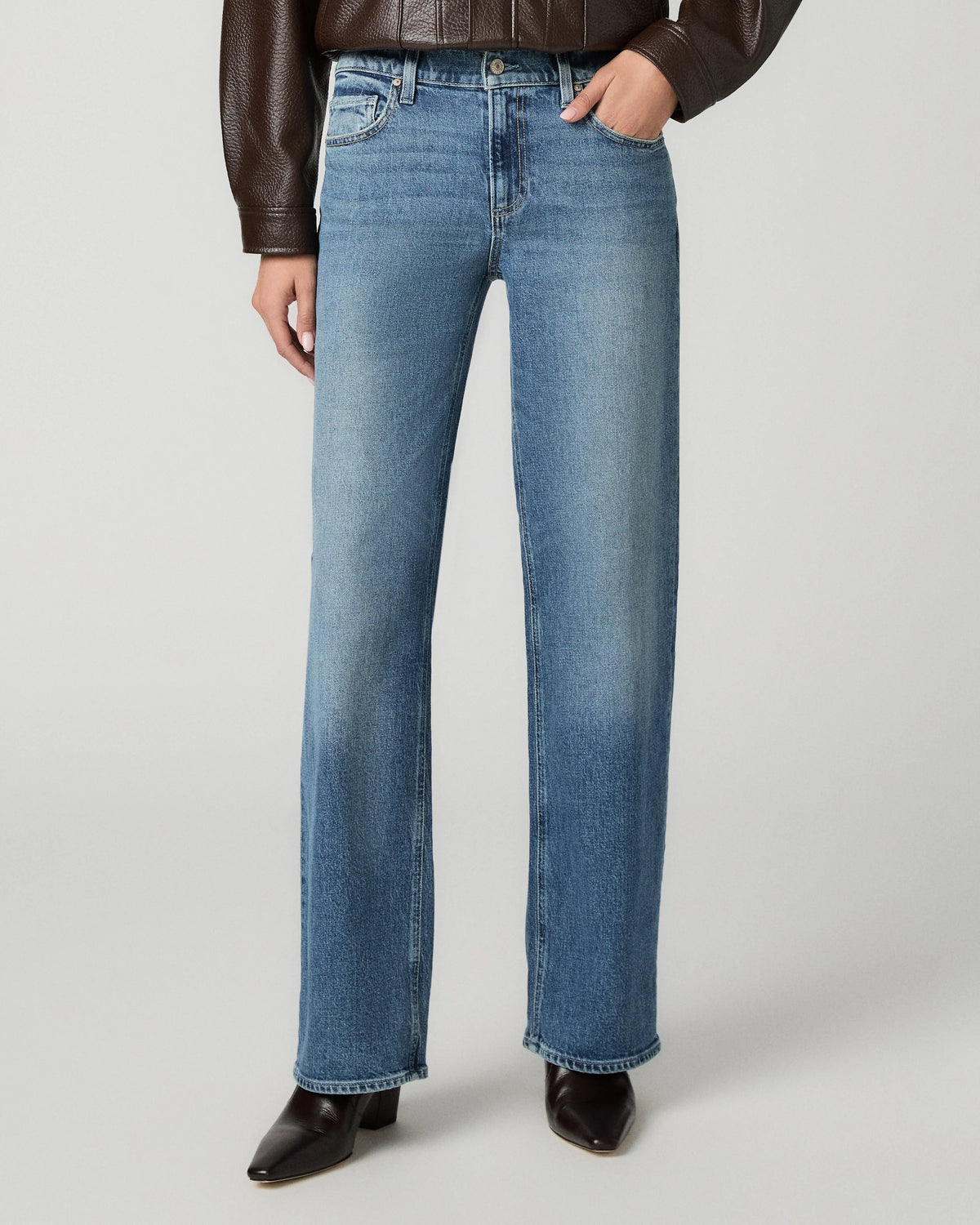A model wearing a pair of medium-wash blue straight-leg jeans, styled with a brown leather jacket and dark brown pointed-toe boots, against a light gray studio background.