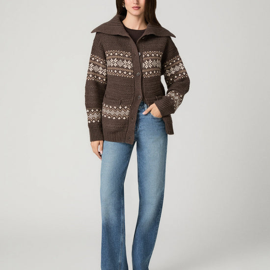 A woman stands against a light gray studio background wearing a brown knit cardigan with cream Fair Isle patterns, blue jeans, and tan shoes.