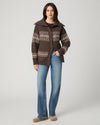 A woman stands against a light gray studio background wearing a brown knit cardigan with cream Fair Isle patterns, blue jeans, and tan shoes.