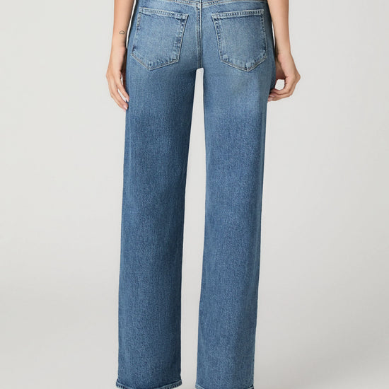 Rear view of a model wearing high-waisted, wide-leg blue denim jeans with a light tan leather patch on the back waistband, paired with black heels against a light gray studio background.