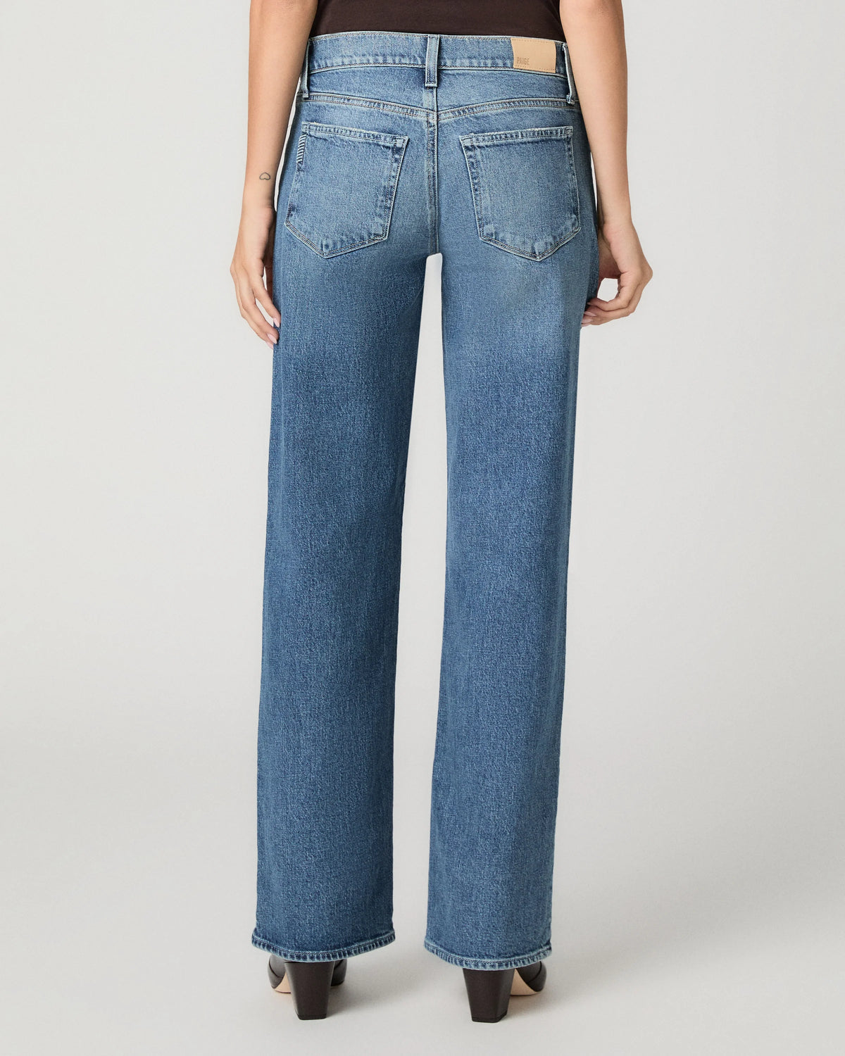 Rear view of a model wearing high-waisted, wide-leg blue denim jeans with a light tan leather patch on the back waistband, paired with black heels against a light gray studio background.