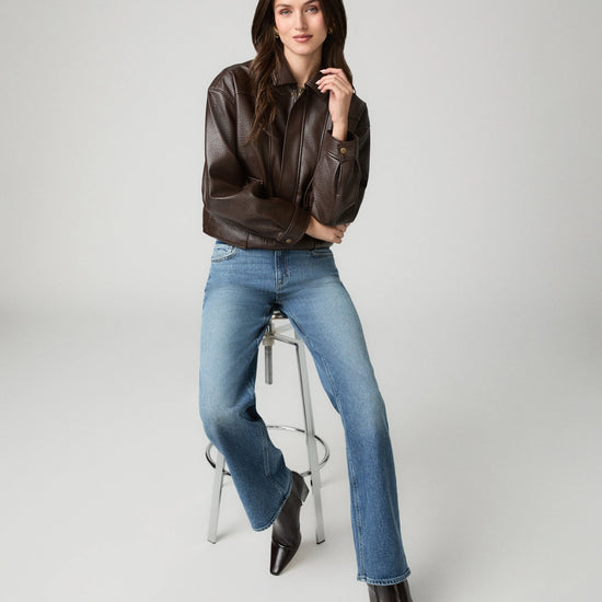 A woman posing on a stool wearing a dark brown leather jacket, blue jeans, and dark brown pointed-toe ankle boots against a light gray studio background.