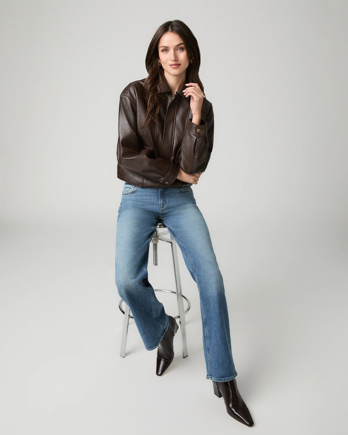 A woman posing on a stool wearing a dark brown leather jacket, blue jeans, and dark brown pointed-toe ankle boots against a light gray studio background.