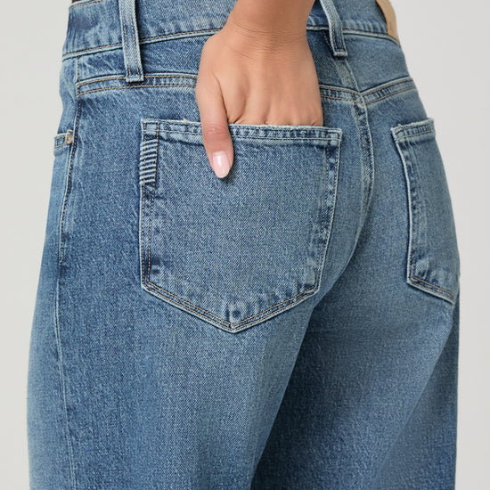 Close-up back view of a person wearing light blue denim jeans, showing the back pockets and waistband with one hand in the pocket, against a plain light gray background.