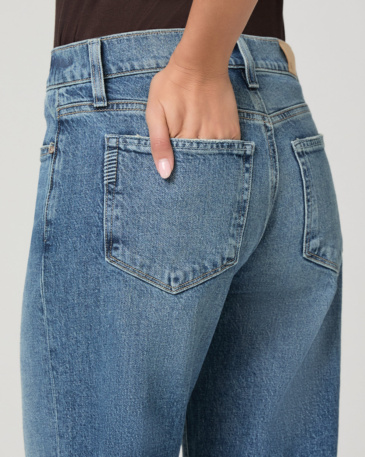 Close-up back view of a person wearing light blue denim jeans, showing the back pockets and waistband with one hand in the pocket, against a plain light gray background.