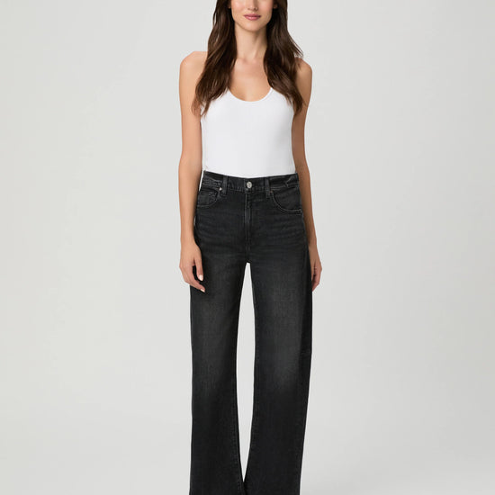 A woman modeling a pair of high-waisted, wide-leg black denim jeans with a white tank top and black sandals, standing against a light gray studio background.