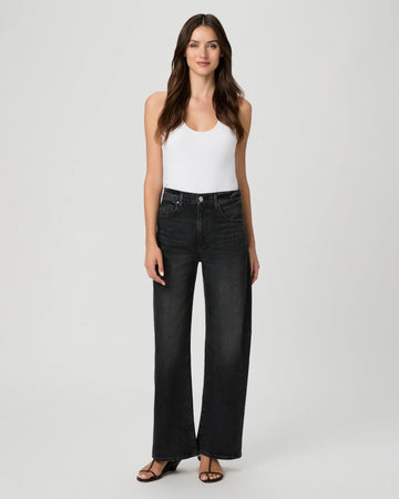 A woman modeling a pair of high-waisted, wide-leg black denim jeans with a white tank top and black sandals, standing against a light gray studio background.