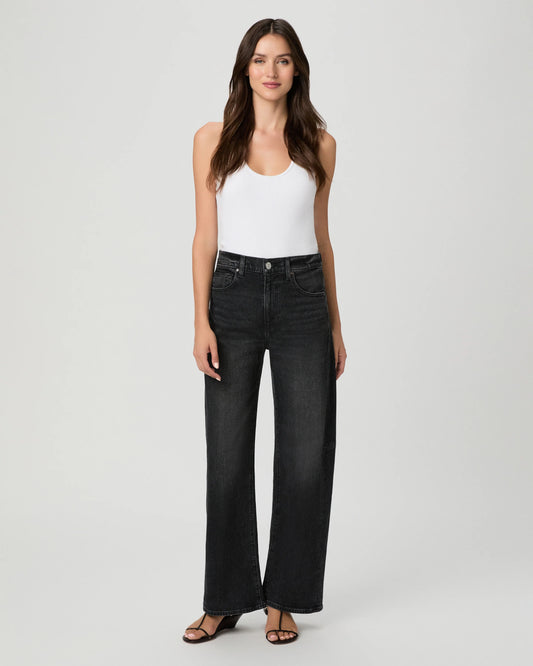 A woman modeling a pair of high-waisted, wide-leg black denim jeans with a white tank top and black sandals, standing against a light gray studio background.