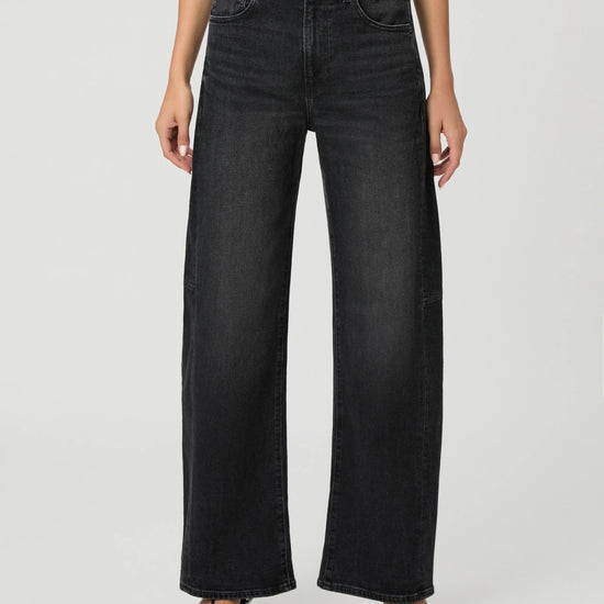 A model wearing a pair of black wide-leg denim jeans with a white top and black sandals, shown from the waist down against a light gray studio background.