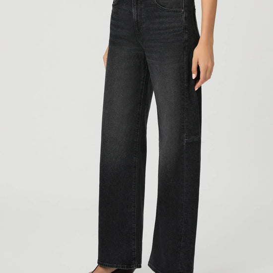 A model wearing a pair of wide-leg black denim jeans with a white top and brown strappy sandals, shown from the waist down against a light gray studio background.