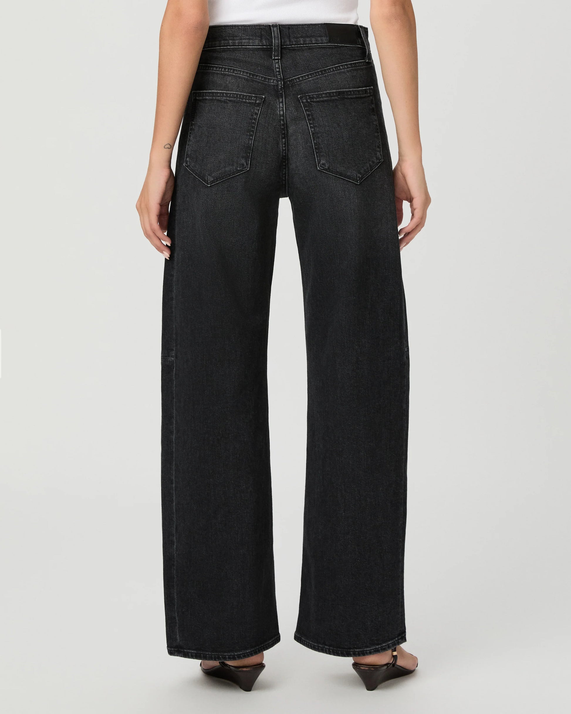 A person shown from the back wearing black wide-leg jeans, a white top, and dark pointed flats against a light gray studio background.