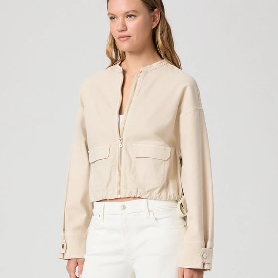 Woman wearing a beige jacket and white pants against a light gray background