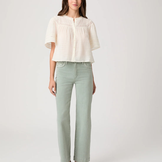 Woman wearing a white blouse by Paige and light green pants on a plain background, showing full outfit 