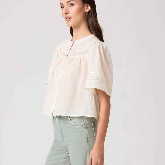 Woman wearing a white blouse by Paige and light green pants on a plain background side view