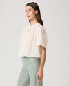 Woman wearing a white blouse by Paige and light green pants on a plain background side view