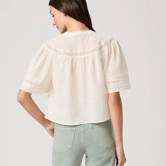 Woman wearing a white blouse by Paige and light green pants on a plain background rear view