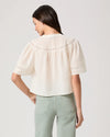 Woman wearing a white blouse by Paige and light green pants on a plain background rear view