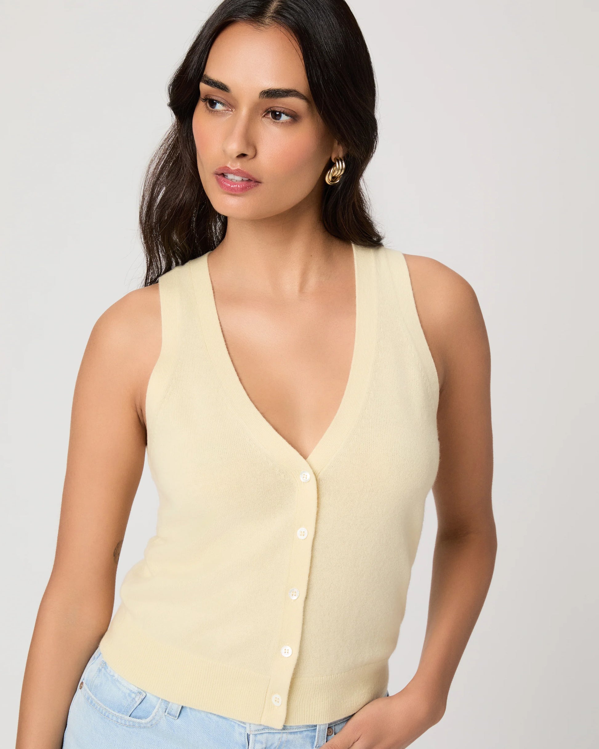 Woman wearing a light yellow sleeveless Paige top and light blue jeans on a plain background close up