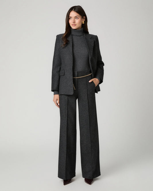 A model stands against a light gray studio background wearing a dark charcoal gray tailored set consisting of a structured turtleneck blazer and matching wide-leg trousers, styled with a thin gold chain belt and dark pointed shoes.