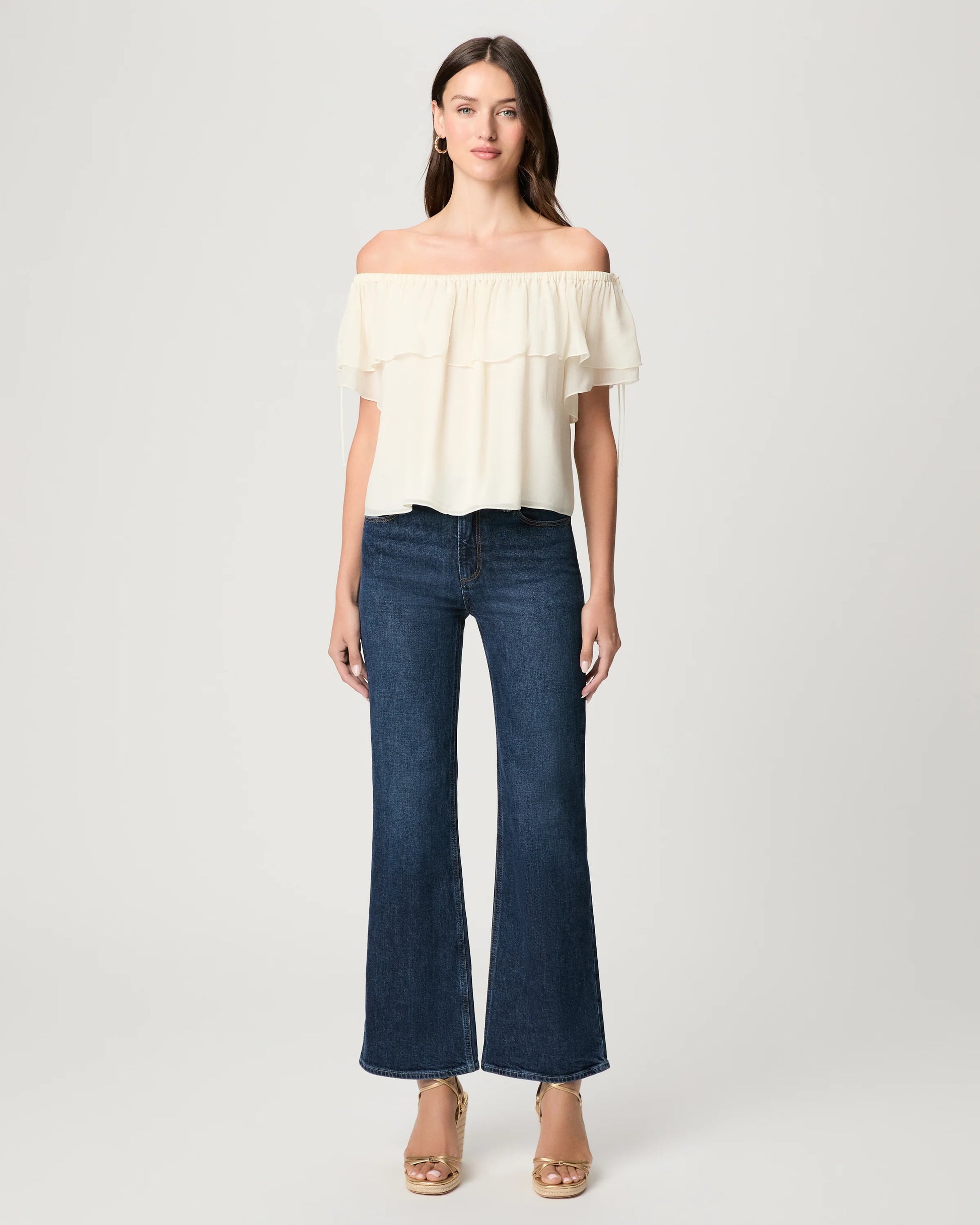 Woman wearing a white off-shoulder top and dark blue jeans on a light gray background