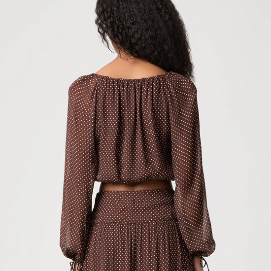 A woman shown from the back wearing a brown off-the-shoulder long-sleeve crop top and matching pleated skirt, both featuring a small white polka dot pattern, against a light gray studio background.