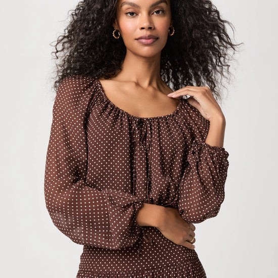 A woman with curly black hair wearing a brown long-sleeve dress with a white polka dot pattern, photographed against a light gray studio background.