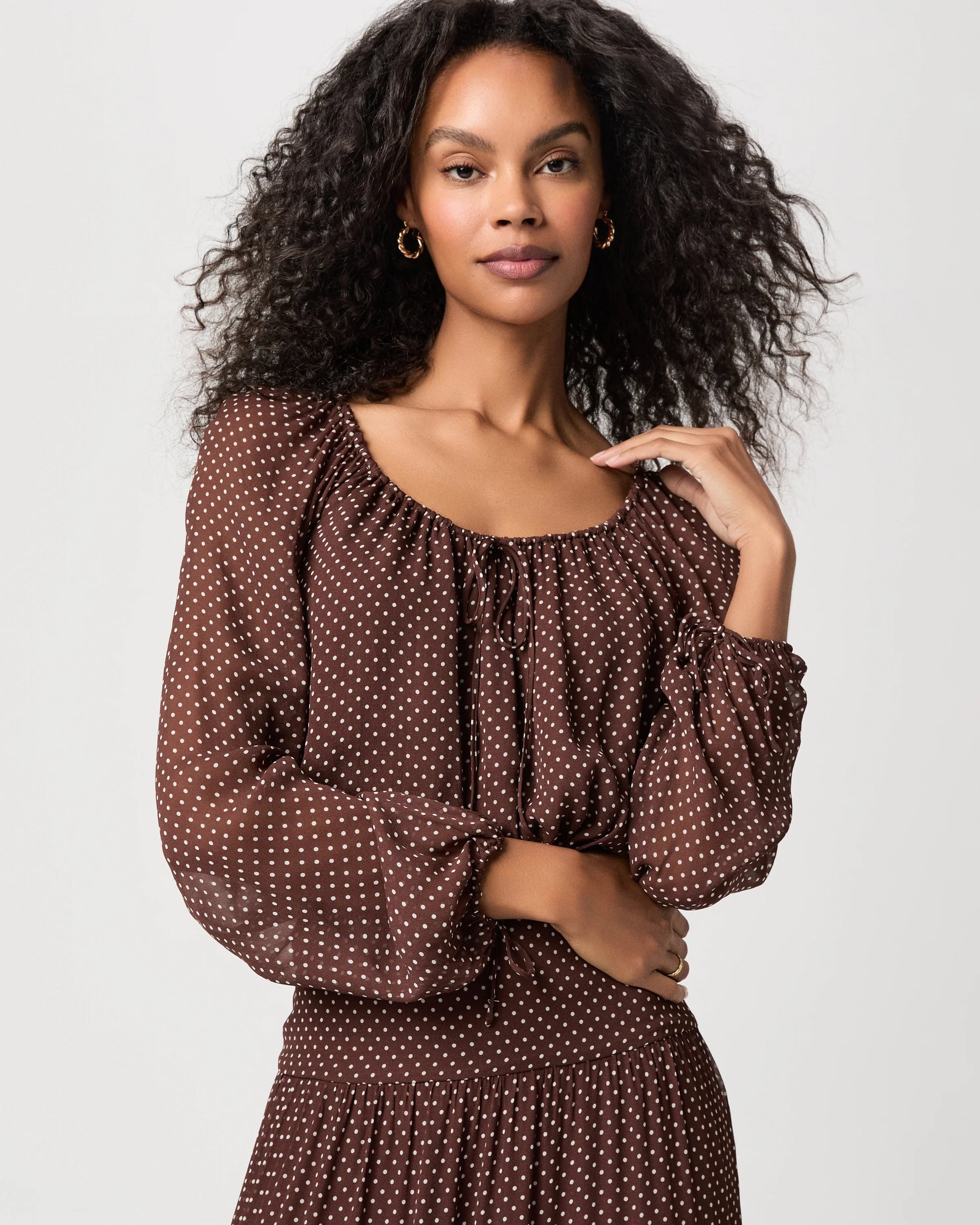 A woman with curly black hair wearing a brown long-sleeve dress with a white polka dot pattern, photographed against a light gray studio background.