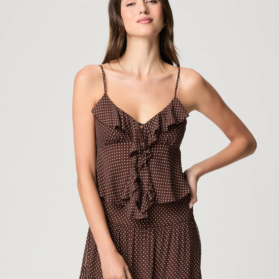 A woman modeling a brown spaghetti-strap dress with a white polka dot pattern and a ruffled tie detail at the bust, set against a plain light gray studio background.