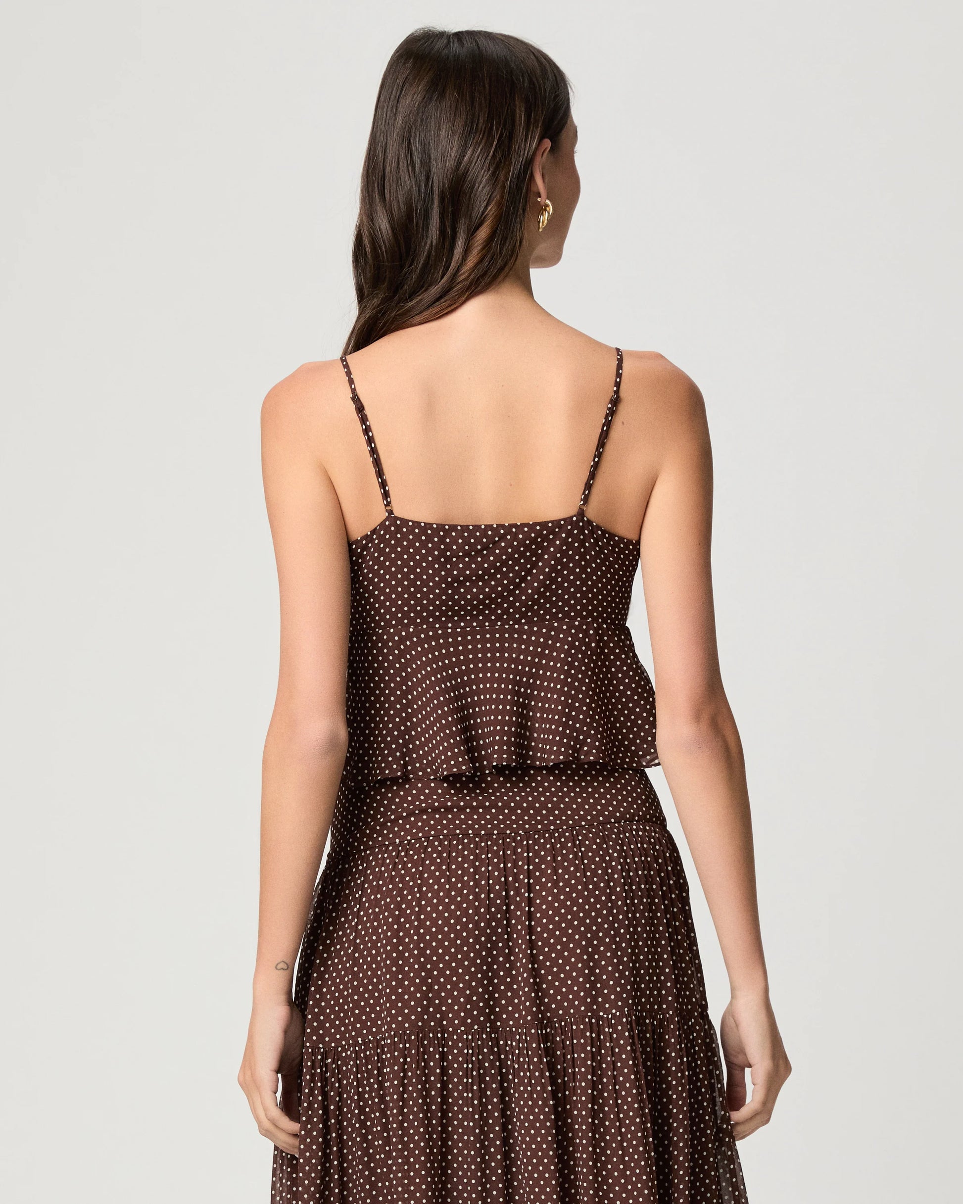A woman shown from the back wearing a dark brown spaghetti-strap dress with a small white polka dot pattern, set against a plain light gray studio background.