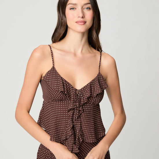 A woman modeling a brown spaghetti-strap dress with a small white polka dot pattern and a ruffled tie detail at the bust, set against a plain light gray studio background.